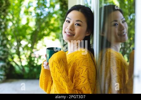 Ritratto di felice donna asiatica indossando maglione giallo in piedi dalla finestra sorridendo e bevendo caffè. Vita domestica, tempo libero e relax. Foto Stock