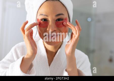 Donna asiatica sorridente che indossa accappatoio e asciugamano guardando in bagno specchio applicare sotto gli occhi patch. Stile di vita, cura di sé, salute e bellezza. Foto Stock