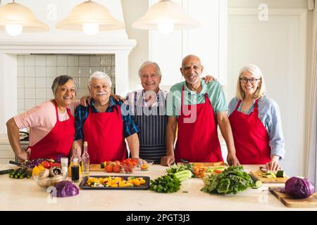 Ritratto di amici anziani felici e diversi che indossano grembiuli in cucina, abbracciando durante la lezione di cucina. Pensione, apprendimento, cucina e sana vita Foto Stock