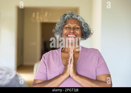 Allegra donna afroamericana anziana sorridente e meditante in preghiera posa a casa, copia spazio. Ritratto, inalterato, stile di vita, pensione, yoga, es Foto Stock