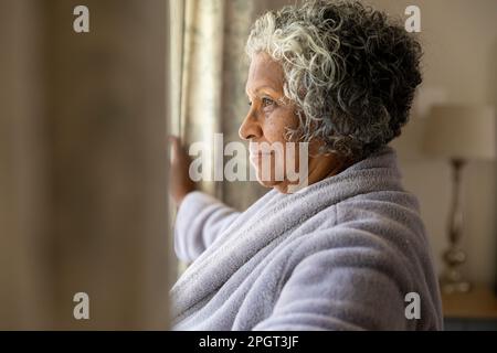 Vista laterale di una donna anziana afroamericana che apre le tende dopo il risveglio al mattino. Copia spazio, inalterato, accappatoio, guardando attraverso la finestra, s Foto Stock