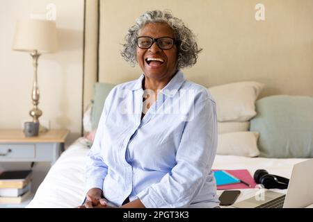Ritratto di afroamericana donna anziana che indossa occhiali ridendo mentre si siede a letto a casa. Copia spazio, allegro, inalterato, lifestyle, relaxa Foto Stock