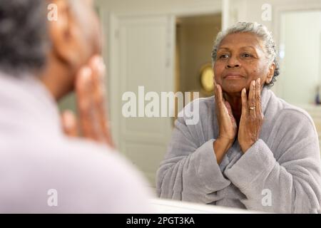 Donna afroamericana anziana con capelli corti grigi che toccano il viso e che guardano in specchio a casa. Inalterato, cura del corpo, cura della pelle, invecchiamento, riflesso Foto Stock