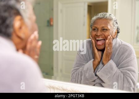 Donna afroamericana anziana che urla allegramente mentre tocca il suo volto e guarda nello specchio. Inalterato, cura del corpo, cura della pelle, invecchiamento, felice, refle Foto Stock