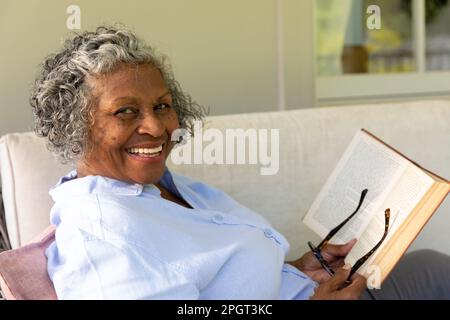 Ritratto di afroamericana senior woman con occhiali e libro sorridente mentre si rilassa sul divano. Copia spazio, inalterato, stile di vita, felice, hobby, hom Foto Stock