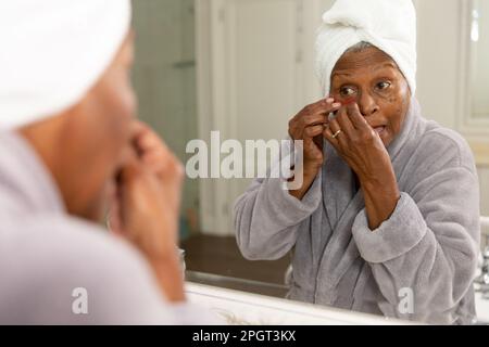 Donna afroamericana anziana vestita di accappatoio guardando in specchio e applicando sotto gli occhi patch. Inalterato, cura del corpo, invecchiamento, riflessione, coccole, Foto Stock