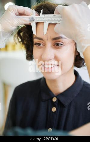 Estetista che utilizza lo strumento di misurazione per la procedura di correzione della fronte in salone di bellezza. Primo piano. Specialista di bellezza che posiziona righello su fronte donna sorridente mentre si effettuano le misurazioni per la correzione delle sopracciglia. Foto Stock