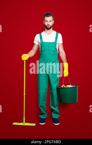 Ritratto integrale di bella e attraente governante con stoppia, acconciatura in uniforme verde, isolato su sfondo grigio, con secchio Foto Stock