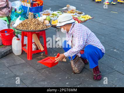 Phnom Penh, Cambogia-Dicembre 23rd 2022:con una paletta e pennello, una donna, la vendita di noci e vari snack di frutta, elimina le briciole e aroun conchiglie di arachidi Foto Stock