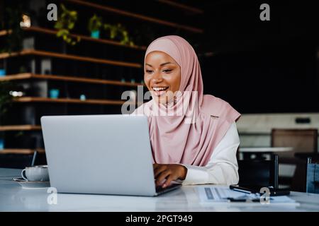 Giovane donna musulmana che indossa il velo sorridendo mentre lavora sul notebook in ufficio Foto Stock