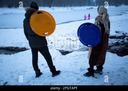 Fine settimana tranquillo. I genitori di Kiev, tenendo grandi piatti di plastica per sciare in discesa sulla neve, guardano i loro bambini camminare lungo la riva del lago ghiacciato Foto Stock