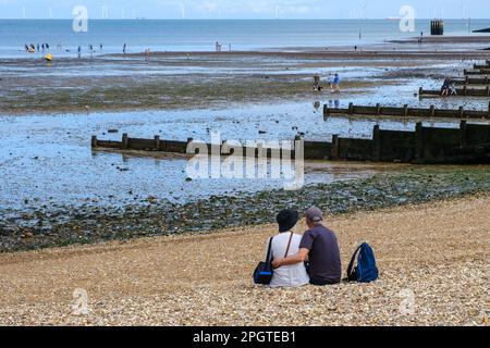 Uomo e donna abbracciano, seduti sulla spiaggia di ciottoli guardare verso il mare e groynes di legno a Whitstable West Beach, nord-est Kent costa, Inghilterra, Regno Unito. Foto Stock