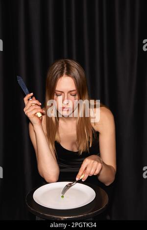 Giovane donna triste con mascara spalmato sulle guance da piangere seduto a tavola e mangiare un pezzo unico pisello sul piatto su sfondo nero Foto Stock