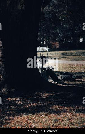 Un giovane soddisfatto seduto in una posizione rilassata in un parco, guardando il suo telefono Foto Stock