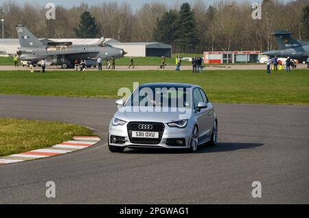 Un'auto che viene guidata ad alta velocità intorno alla pista di prova di Bruntingthorpe Proving Ground, sul sito dell'Aerodromo di Bruntingthorpe con jet vintage oltre Foto Stock