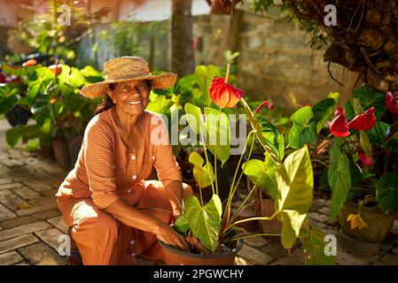 Senior femmina contadina indiana che piantano fiori sorridendo felicemente. Anziana ragazza dello Sri Lanka allegra seduta nel suo giardino curandosi delle piante in fiore Foto Stock