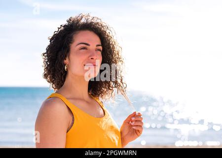 Giovane donna caucasica graziosa che tiene un filo dei suoi capelli ricci con le dita sulla spiaggia mentre sta guardando in su. Foto Stock