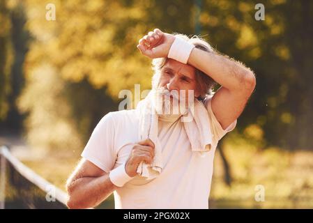 Uomo elegante e anziano in camicia bianca e pantaloncini sportivi neri sul campo da tennis Foto Stock
