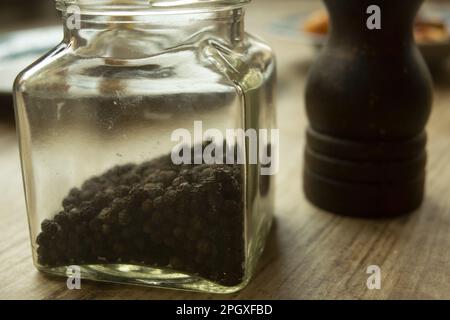 grani di pepe nero in un vaso di vetro con un tritacarne sullo sfondo Foto Stock