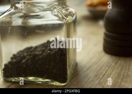 grani di pepe nero in un vaso di vetro con un tritacarne sullo sfondo Foto Stock