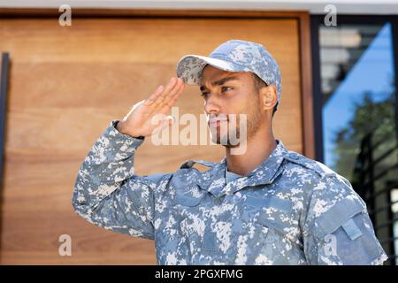 Soldato biraciale maschio che indossa uniforme e cappuccio, saluta fuori casa Foto Stock