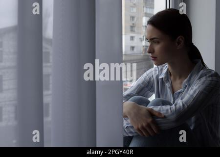 Giovane donna malinconica che guarda fuori dalla finestra al chiuso, spazio per il testo. Concetto di solitudine Foto Stock