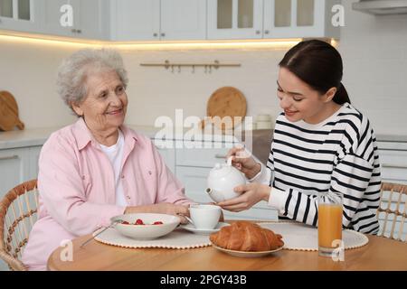 Giovane assistente che serve la colazione per le donne anziane al tavolo in cucina. Assistenza domiciliare Foto Stock