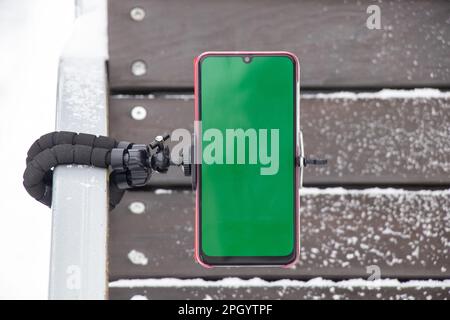 un telefono cellulare con schermo verde fissato su un supporto flessibile sulla ringhiera di una panca all'aperto in inverno Foto Stock