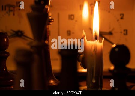 pezzi di scacchi in piedi su una scacchiera e orologio e accanto a una candela che brucia al buio, giocando a scacchi Foto Stock