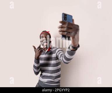 Giovane uomo africano allegro, con un maglione a righe grigie, un volto divertente e un gesto d'amore rock 'n' roll mentre si prende un selfie smartphone. Foto Stock