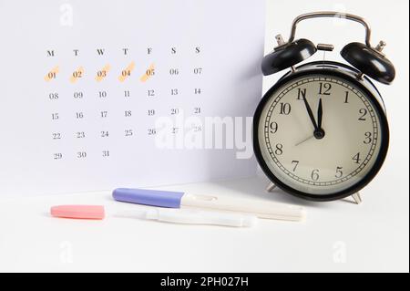 Test di gravidanza a getto d'inchiostro positivo, orologio di allarme retro nero e calendario bianco con le date dell'ultima mestruazione marcata Foto Stock