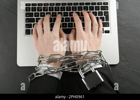 Dipendenza da Internet. Uomo che digita sul computer portatile con le mani incatenate al tavolo nero, vista dall'alto Foto Stock