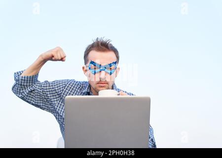 Uomo caucasico in camicia da notte e maschera blu supereroe con laptop e gesto di forza. Fists pompati o superpotenza, concetto serio di emozione. Immagini di alta qualità Foto Stock