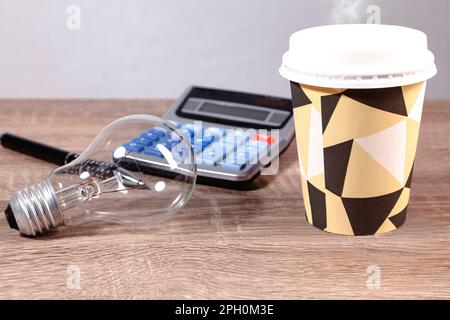 Sopra un tavolo di legno ci sono una lampadina, fumo caffè da andare, penna e calcolatrice. Foto Stock