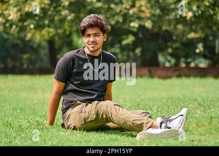 Sorridente ritratto di un uomo indiano in t shirt nera e catena del collo d'argento seduto su prato verde in fondo al parco pubblico, indù attraente portatore maschile Foto Stock