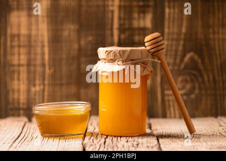 Miele in un vaso di vetro chiuso e una ciotola, cucchiaia di legno su uno sfondo di legno. Composizione di vasetti e cucchiai di miele. Prodotti biologici. Foto Stock