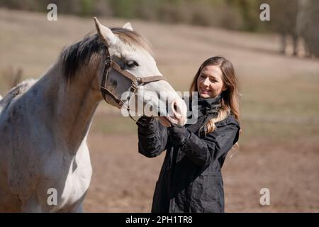 Giovane donna in piedi accanto al cavallo arabo bianco, fissando bridle, il sole splende su di loro, dettaglio primo piano Foto Stock
