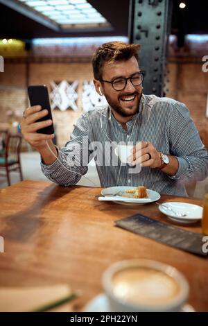 Giovane gioioso che si diverte a gustare un caffè al bar, ascoltando musica, sorridendo. Un uomo elegante e bello che si è sempre bene. Stile di vita, concetto di lisiure. Foto Stock