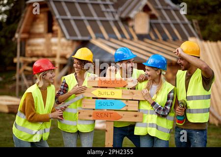 Un gruppo di giovani e allegri costruttori, sia femminili che maschili, amano posare per una foto in un luogo di costruzione di cottage in una bella giornata. Costruzione, buil Foto Stock