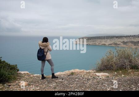 Donna turistica non riconosciuta che si trova ai margini della collina e che si gode il panorama di una costa rocciosa Foto Stock