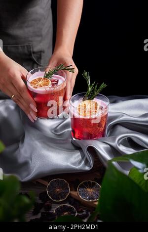 Cocktail di rum e mezcal con succo di lime e ciliegie guarnito con frutta secca e rosmarino verde posto da due mani femminili, piante Foto Stock