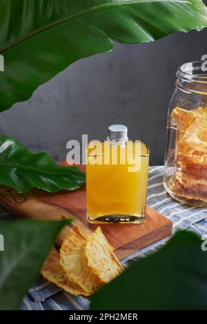Vasetto di vetro con speciale cocktail di ananas, rum e mezcal con peperoncino e piante di ananas guarniti con frutta secca in una cucina con piano Foto Stock