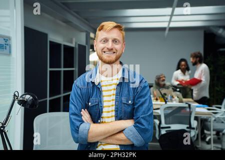 Giovane programmatore con capelli rossi o uomo d'affari in un moderno ufficio guardando fotocamera e sorrisi. Foto Stock