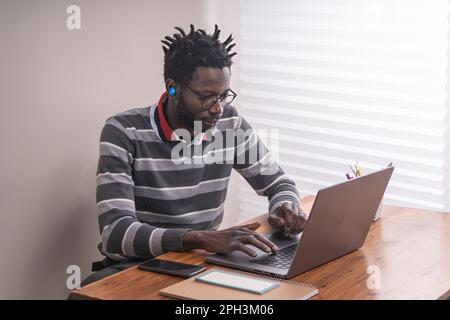 Studente afro-americano che indossa occhiali e cuffie intrauricolari wireless, digitando su una tastiera durante una chiamata. Seduti ad una scrivania di legno senza Foto Stock