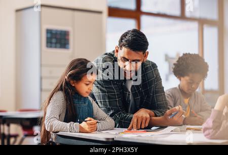 Uomo che guida i bambini della scuola elementare. Insegnante che mostra ai bambini come disegnare in una classe. Educatore maschile aiuta una giovane ragazza su come completare un arte ta Foto Stock