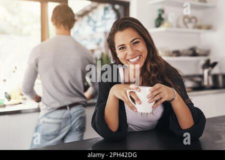 Non si può dire il caffè senza un sorriso. una giovane donna che ha un caffè in cucina con il suo ragazzo in piedi sullo sfondo. Foto Stock