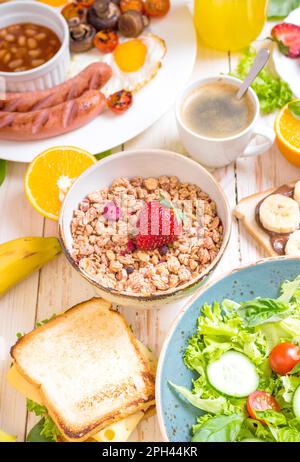 Assortimento di scelte per la colazione. Granola con fragola in una ciotola e salsicce, uova fritte, pancetta, insalata, panino al formaggio, pancake, crema di cioccolato Foto Stock