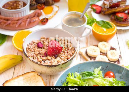 Assortimento di scelte per la colazione. Granola con fragola in una ciotola e salsicce, uova fritte, pancetta, insalata, panino al formaggio, pancake, crema di cioccolato Foto Stock