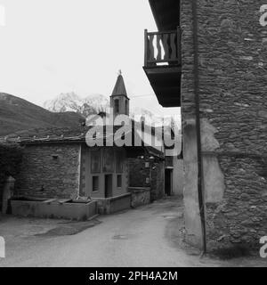 Villaggio di Nus con una chiesa e casa con balcone. Valle d'Aosta, Italia. Acqua attraverso il primo piano e le alpi innevate dietro. Monocromatico Foto Stock