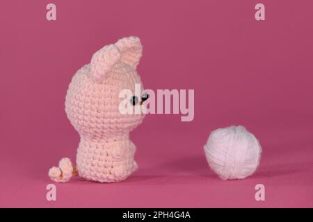 Amigurumi un carino piccolo gattino seduto su sfondo rosa accanto alla palla bianca di filo. Soffice giocattolo fai da te in cotone naturale e lana. Gatto uncinetto, Foto Stock